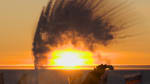 A person in shadow as they are throwing boiling water into the air, creating an arc of ice crystals, in front of the sunset at the ceremonial Pole.