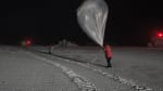 Winterover in red parka, against dark black sky, holding weather balloon for launching.