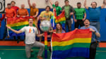 Group photo in the South Pole gym, with people in t-shirts the colors of the rainbow, and with arms up and holding rainbow flags.