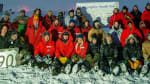 South Pole winterover group photo under a star-filled bluish sky with a horizontal streak of green auroras and a vertical green laser beam shooting up into the sky.