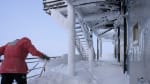 Winterover in red parka shoveling thick snow on deck of the IceCube Lab.