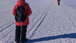 A winterover in red parka shown from behind as they film another winterover in red parka who is walking away from them toward the South Pole Telescope in the distance.