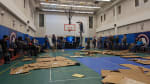 People lined up along walls of gymnasium, lots of flattened carboard strewn about the floor.