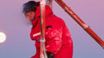 Person in red parka seen sitting on metal tower structure at the South Pole during sunset, beautiful pinks and purples in sky.