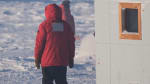 A twin otter, blurry in the far distance, with the backs of two onlooking winterovers in red parkas in the foreground.