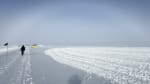 A white rainbow (or fogbow) on the horizon at the South Pole, with a person walking ahead in the distance.