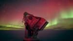 Beautiful green and red auroras stretching upward from low on the horizon at the South Pole, with the South Pole Telescope barely lit in red in the foreground.