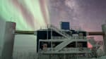 The IceCube Lab with a pseudo-split-screen background of Milky Way and stars off to the right and auroras off to the left.