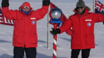 Two men in red parkas standing at the ceremonial South Pole