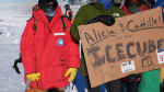 Group in parkas holding large cardboard sign out on the ice with parked plane behind them.