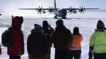 Group of people seen from behind in the foreground, large propellor plane approaching on ice in the background.