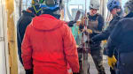 Group in parkas and hardhats lowering drill head down into hole.