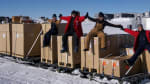 Group photo of four people sitting on top of large boxes, with arms stretched out sideways linking hands.