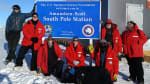 Group photo of eight people standing around the South Pole station sign.
