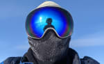 Close-up of winterover’s face covered by mask and googles, which are reflecting the parka-wearing photographer and the sun behind him.