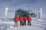 Four winterovers in red parkas pose on the ice in front of the IceCube Lab.