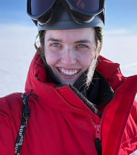 A woman wearing a red jacket and ski googles smiling.