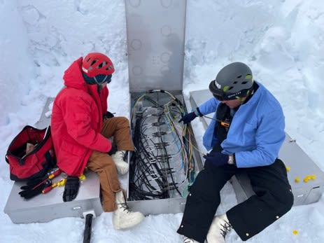 Two people seated on ground around an open junction box.