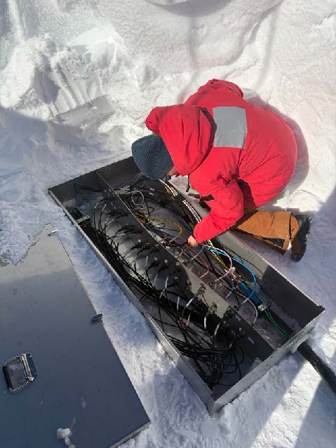 A person in red parka crouched over inspecting connections in a junction box out on the ice.