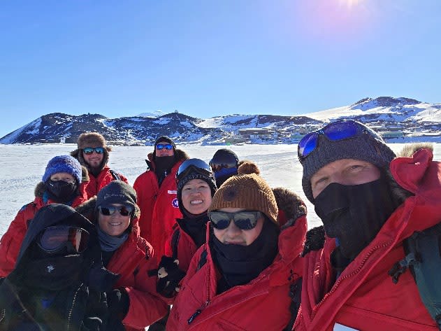 Group in red parkas out on the ice, smiling for photo, mountains in distant background