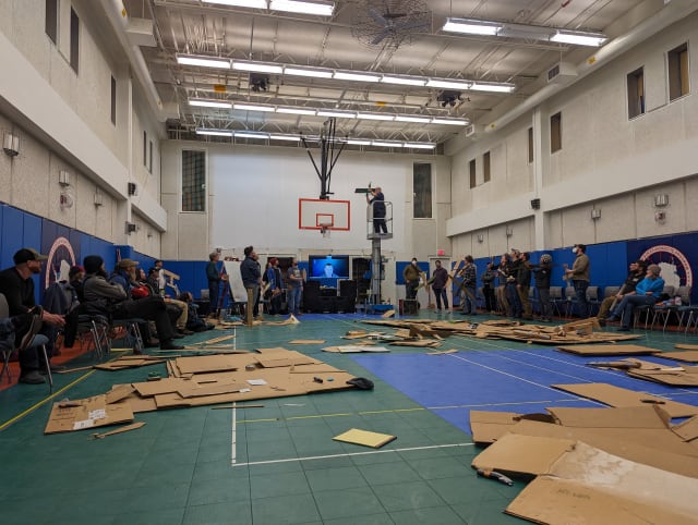 People lined up along walls of gymnasium, lots of flattened carboard strewn about the floor.