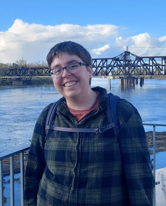 A person smiling in front of a bridge over water.