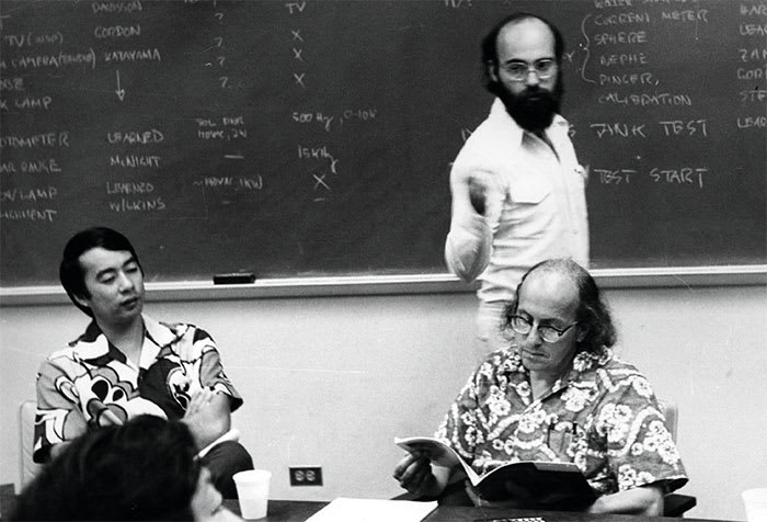 Two men in tropical shirts are seated at a table; the one on the right has glasses and is reading a pamphlet. Behind him, a man stands at a chalkboard.