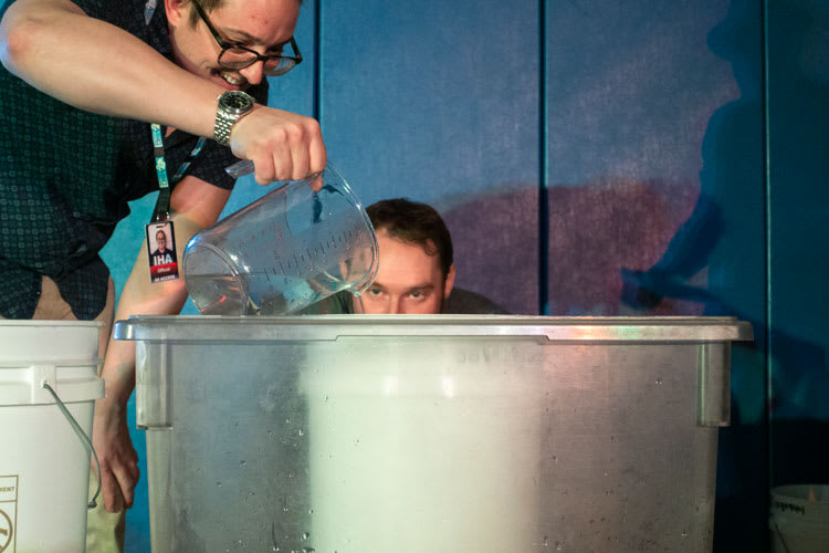 Someone pouring water from clear pitcher into large container, with partial view of another person’s face as they watch.