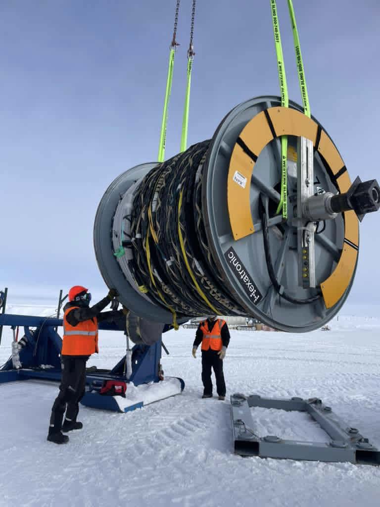 Two people working on large cable spool on crane about 4 feet off the ground.