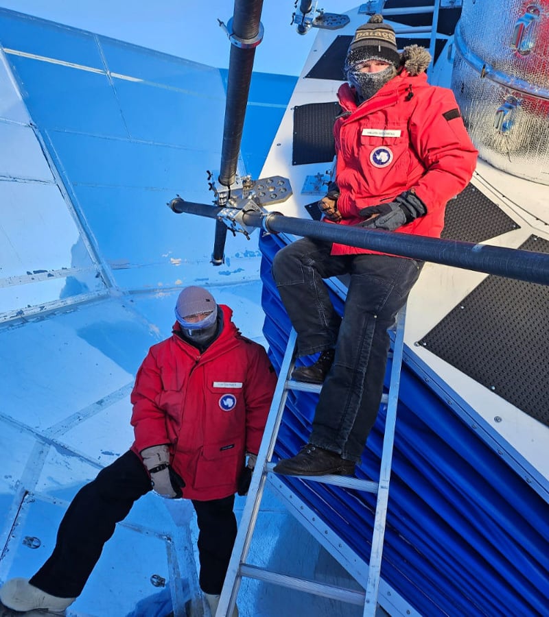 Two winterovers in red parkas, one on ladder, other standing beside, in the BICEP array at the South Pole.