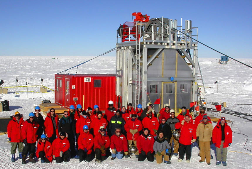 A group photo from the first season of IceCube construction at the South Pole. The group is in front of one of the towers used for drilling holes in the ice.