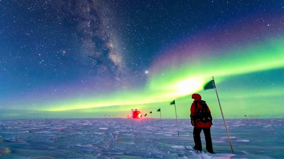 A winterover at the South Pole walking along a flag line that leads to the IceCube Laboratory. The night sky shows the Galactic plane and aurora australis