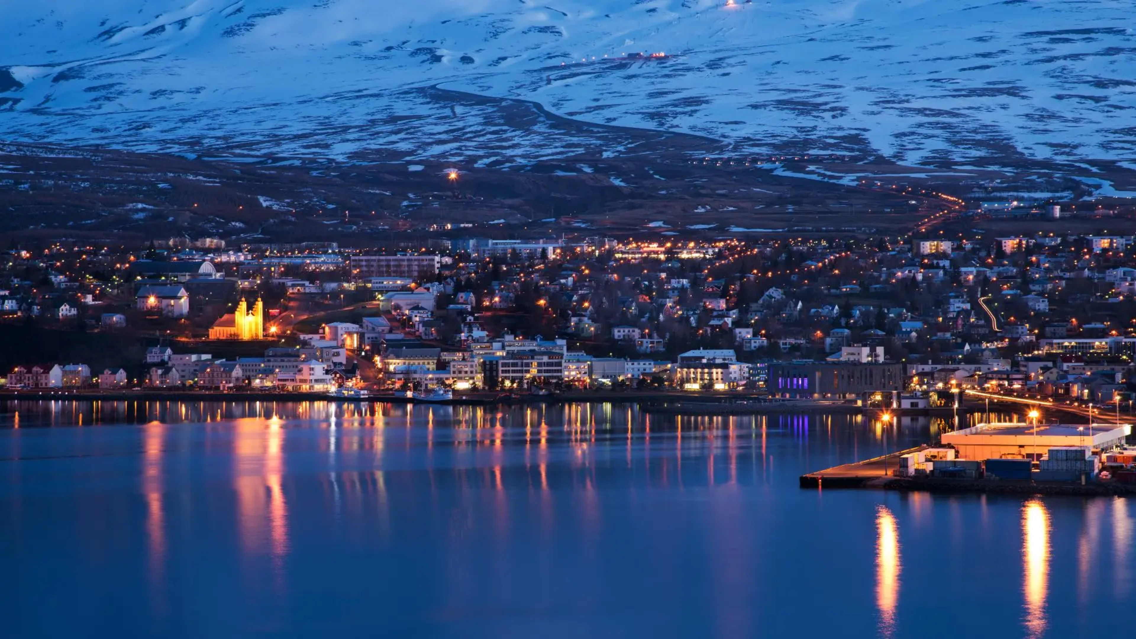 city by snowy mountain at night with house and city lights reflecting in water