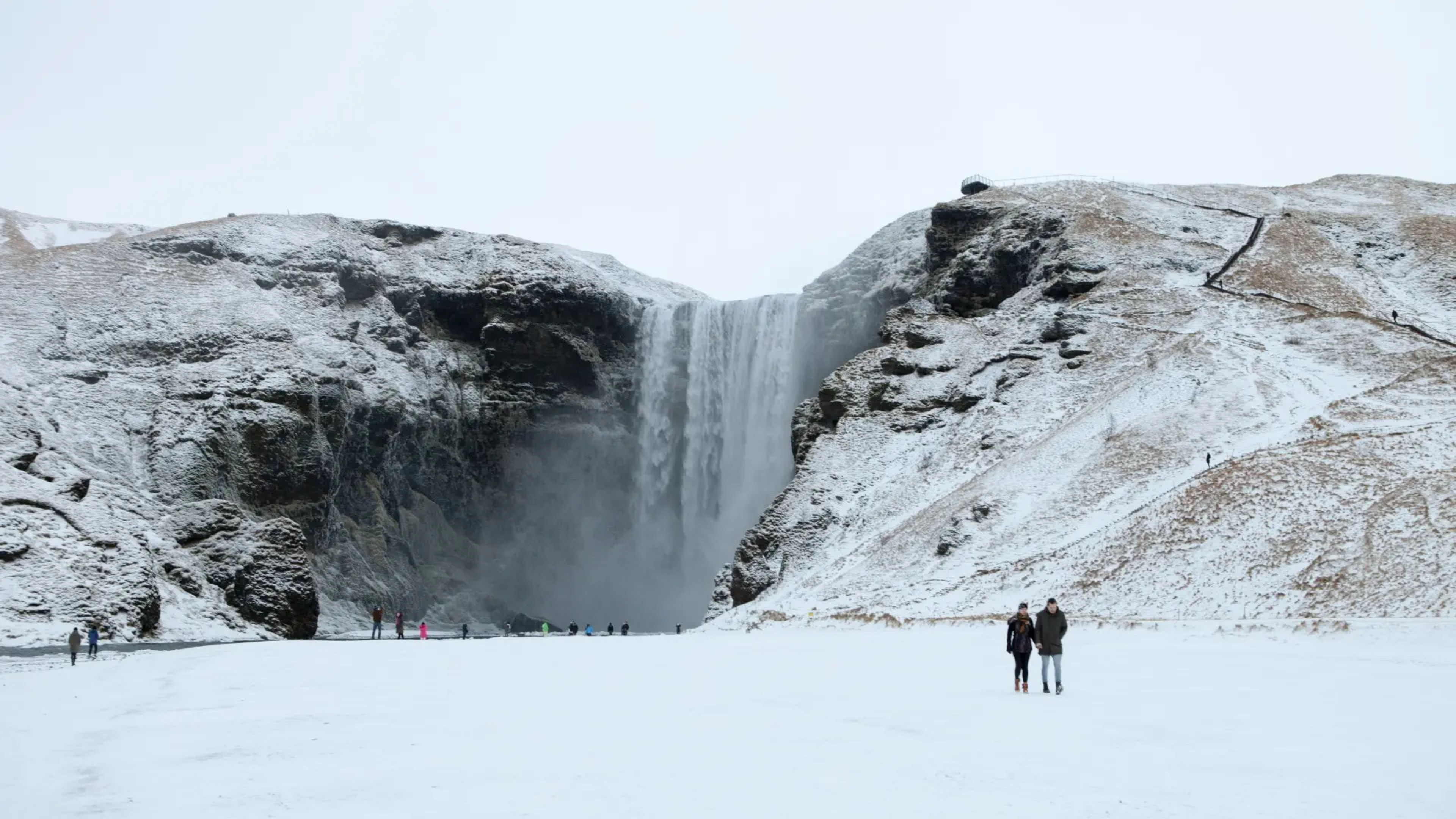 couple walking along snowy plain to a tall waterfall and rock face