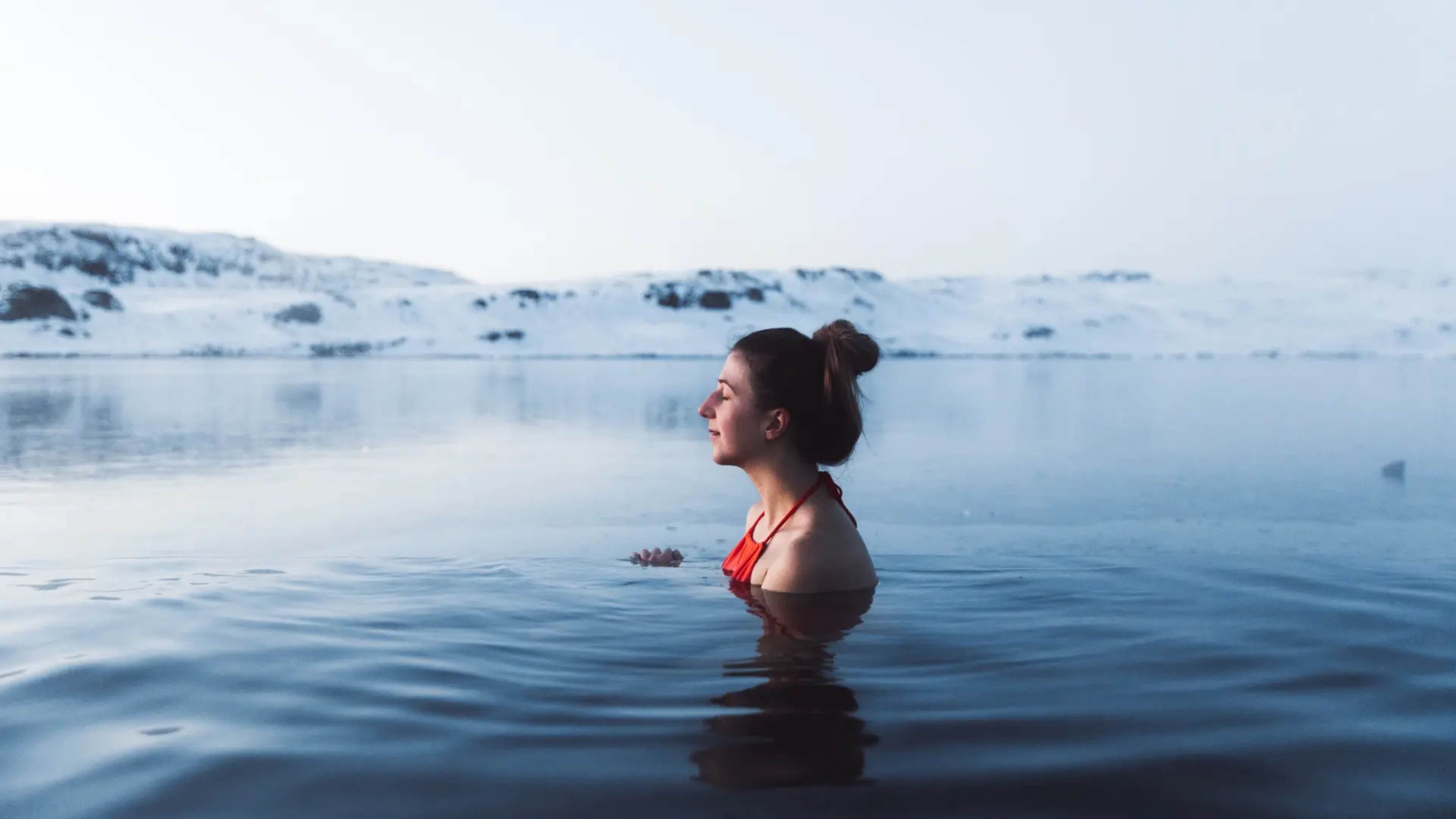 A woman with her eyes closed relaxing in water with snowy hills in background