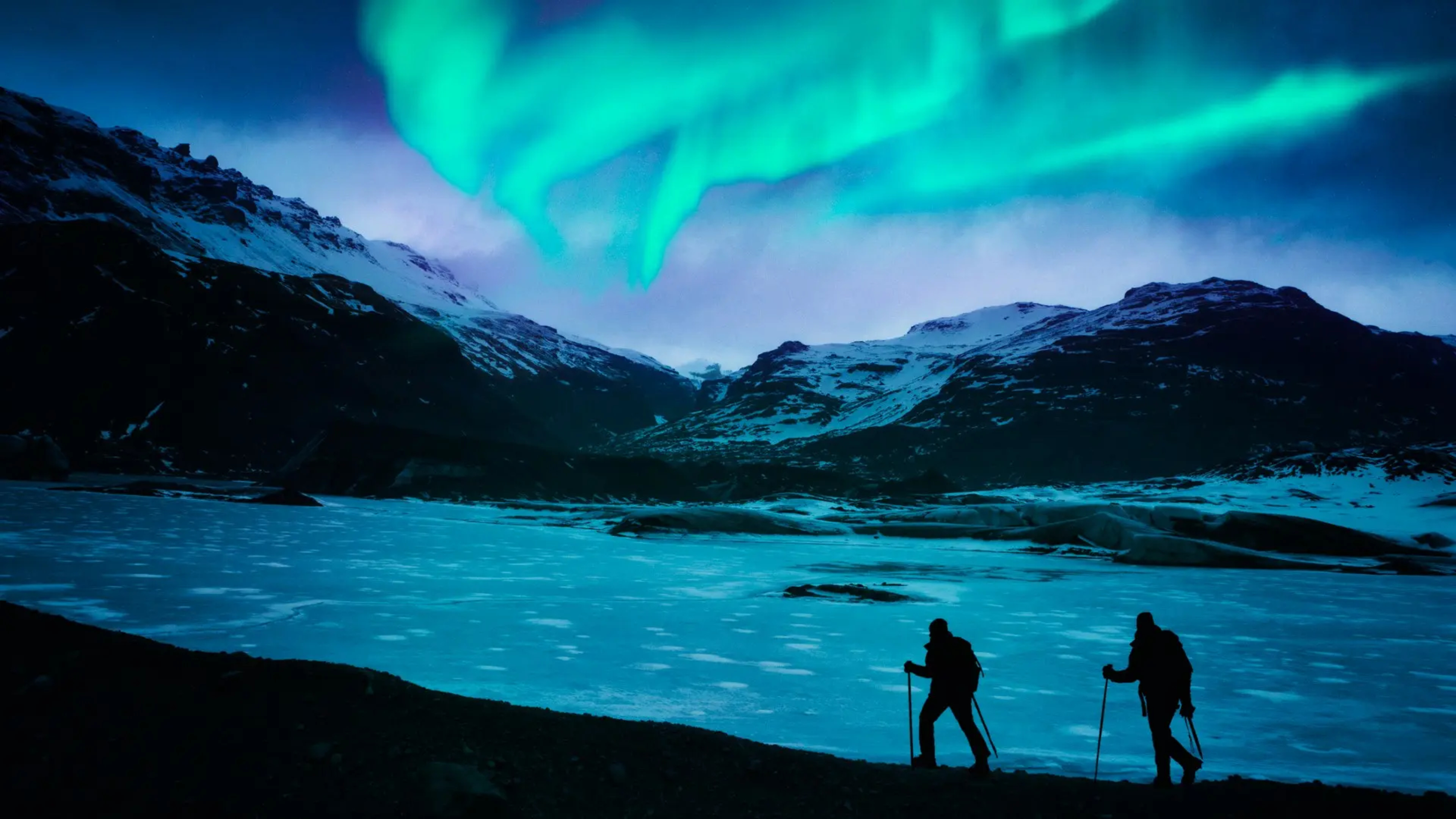 two people hiking along lagoon at night with green northern lights in the sky