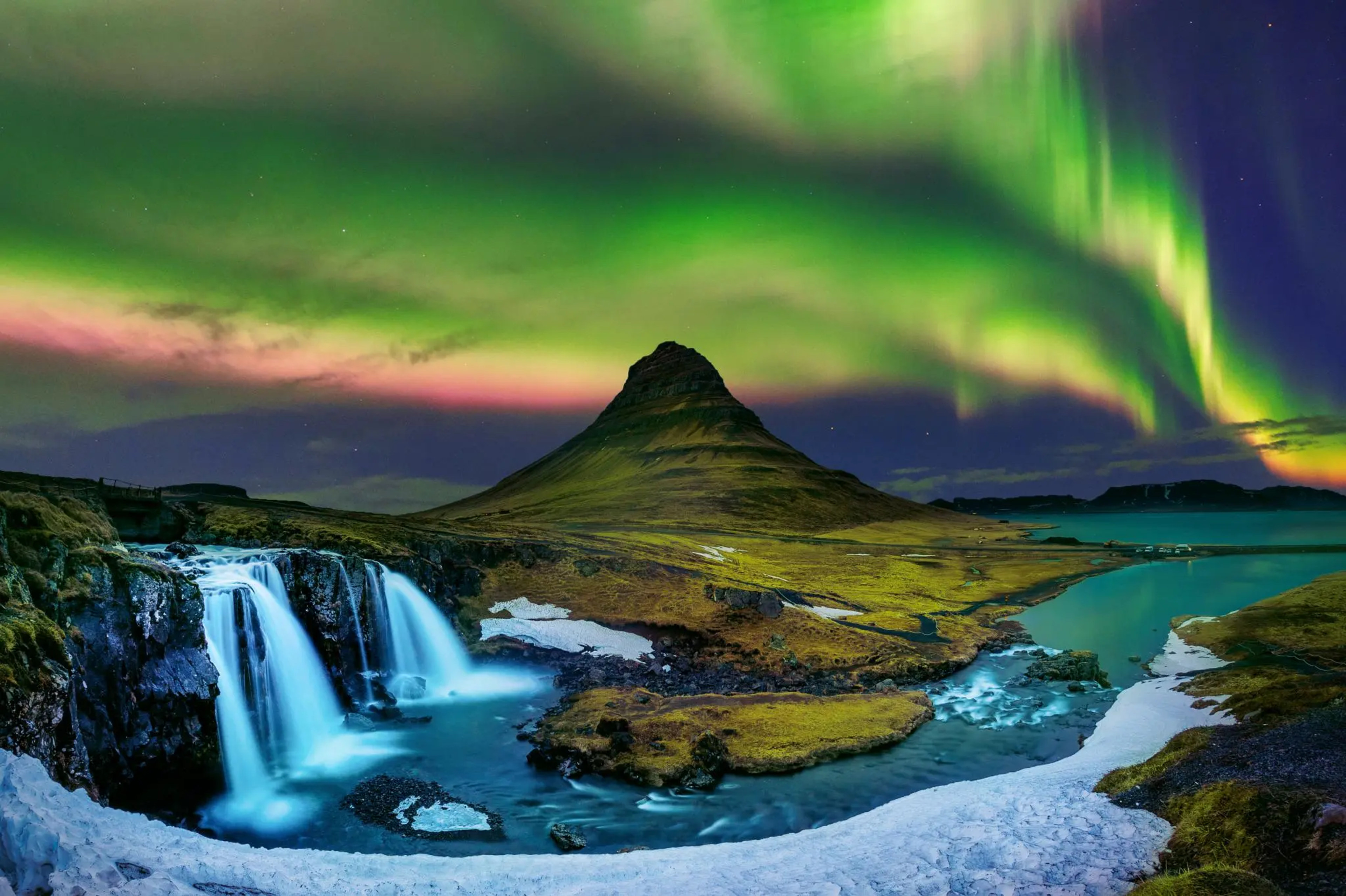 Green and pink northern lights in the sky above pointy mountain and waterfall