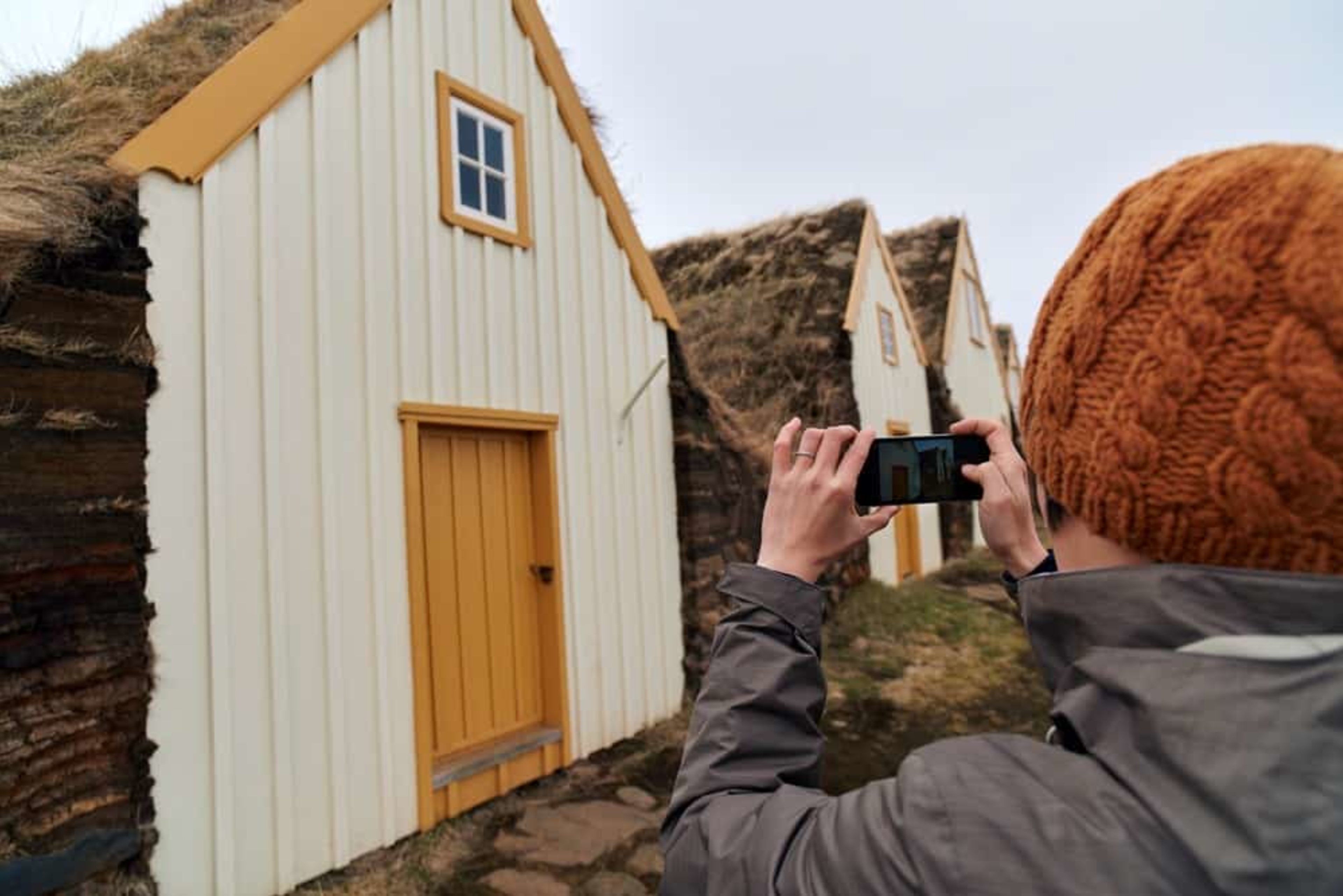 person with orange hat taking a photo on their phone of row of white turf houses