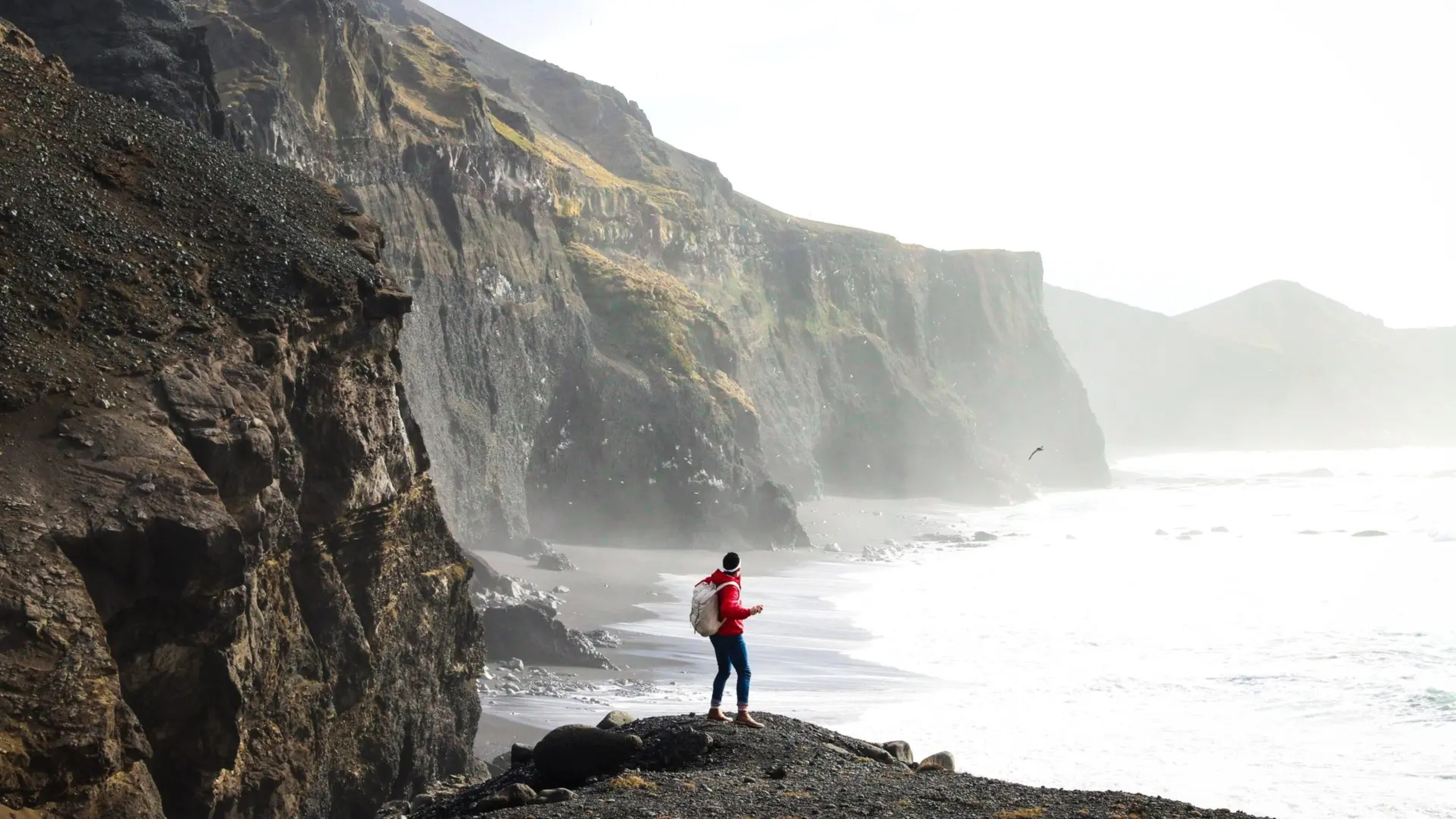person standing by a cliff with rock face above and sea below