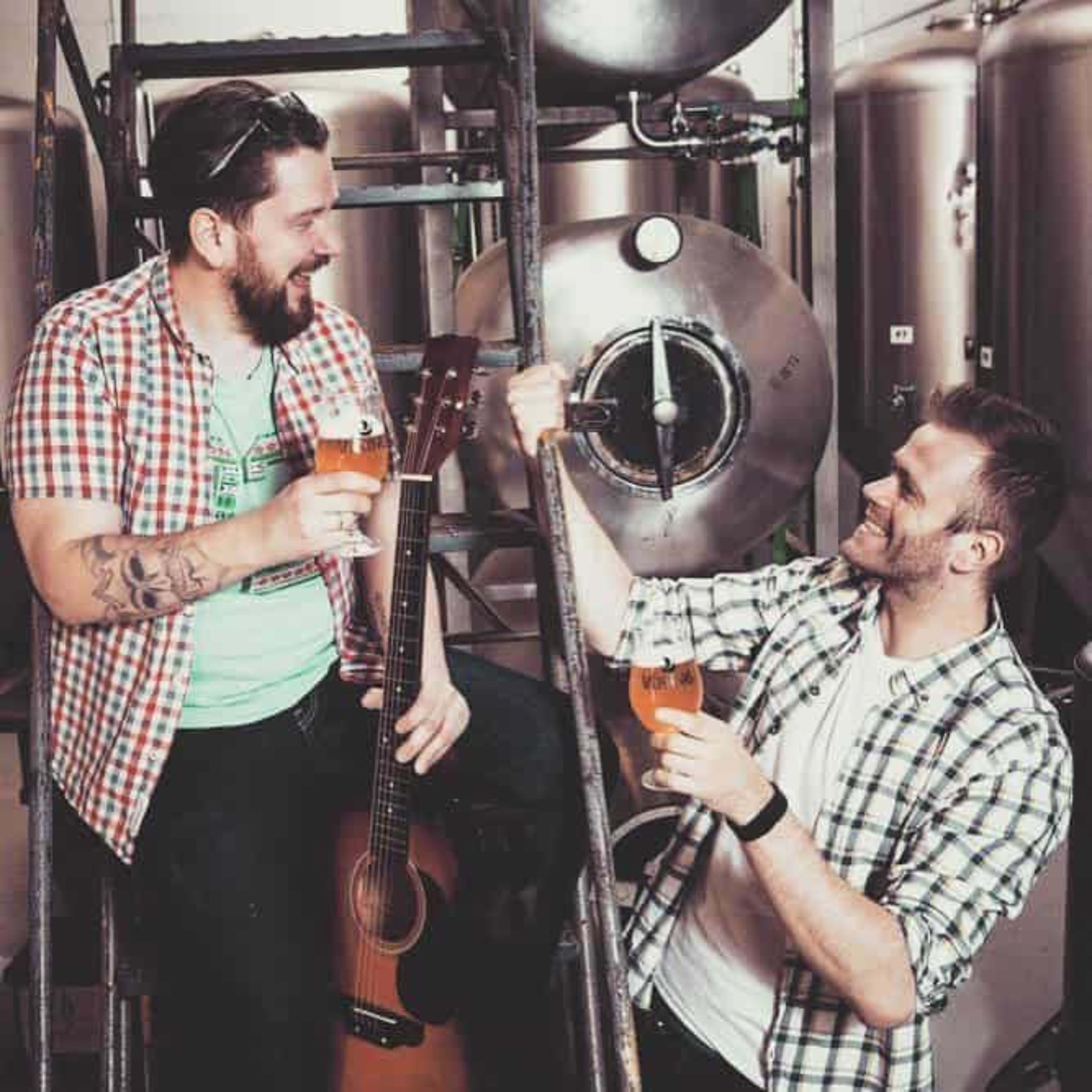 2 men with beer glasses in a brewery, one is holding a guitar