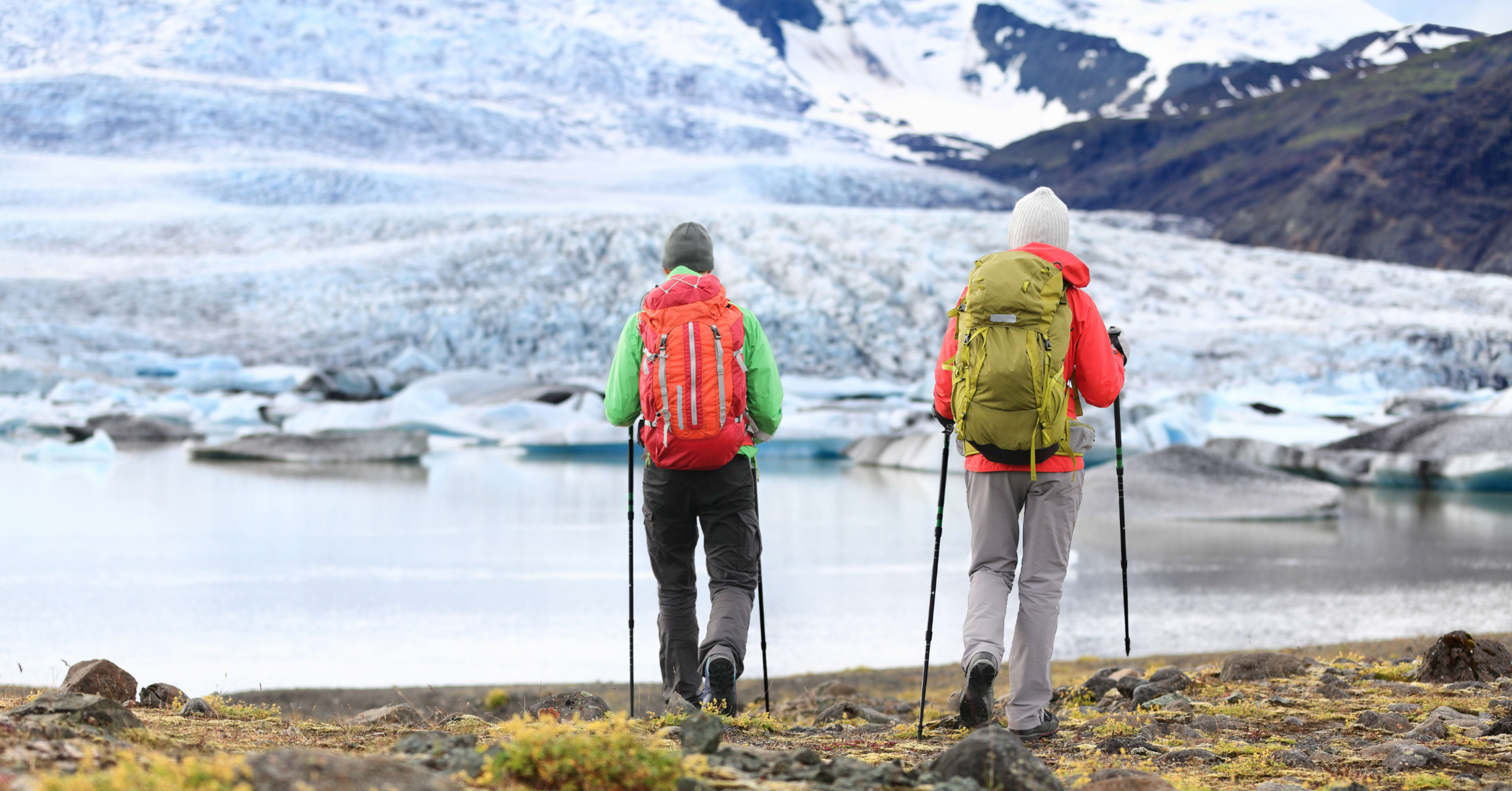Two hikers with trekking poles and colorful backpacks walking toward a glacier lagoon surrounded by ice and snowy peaks.