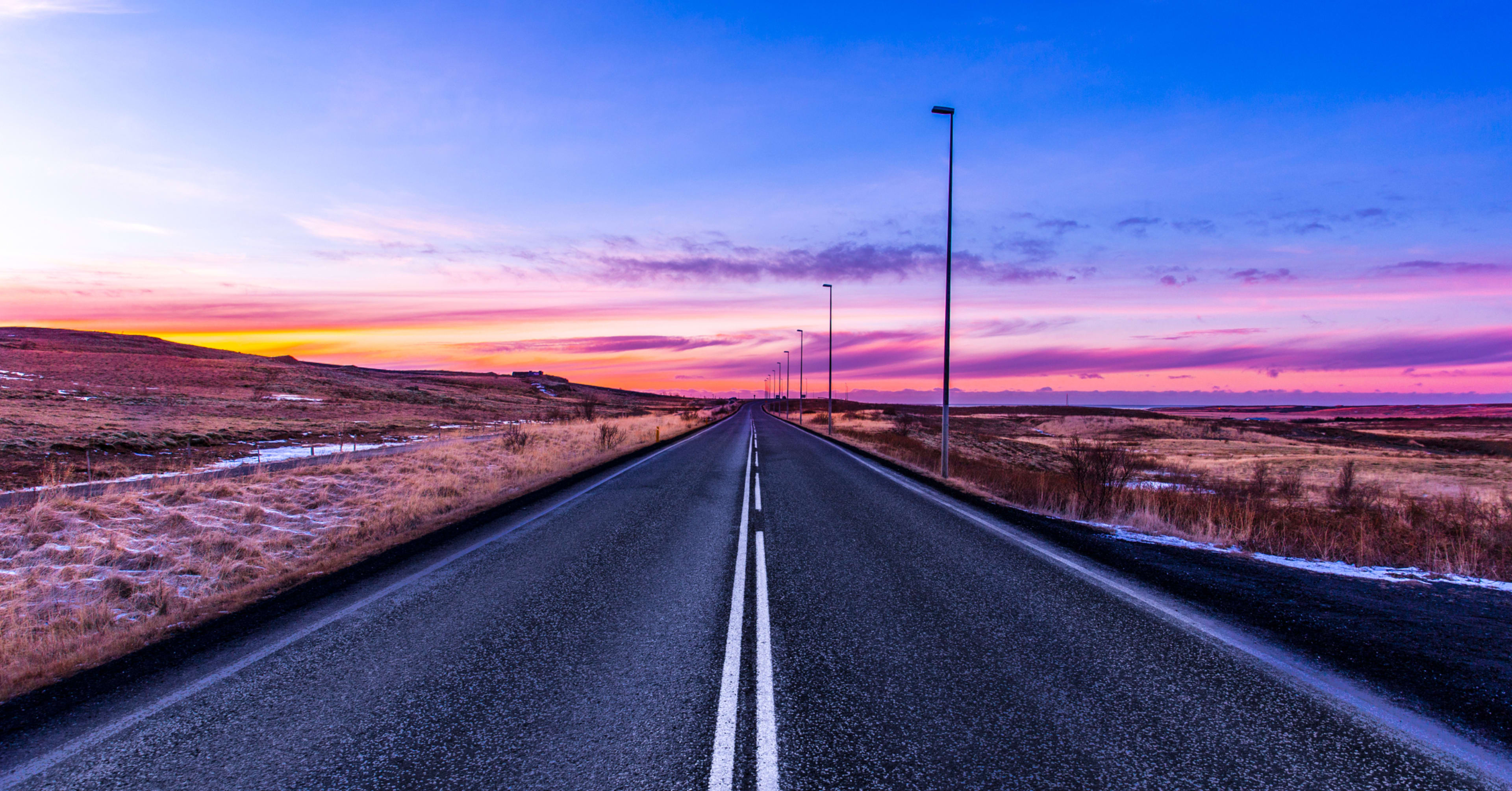 Straight road in rural Iceland at sunrise with pink and purple skies, streetlights lining both sides, and frosty grass on the shoulders.