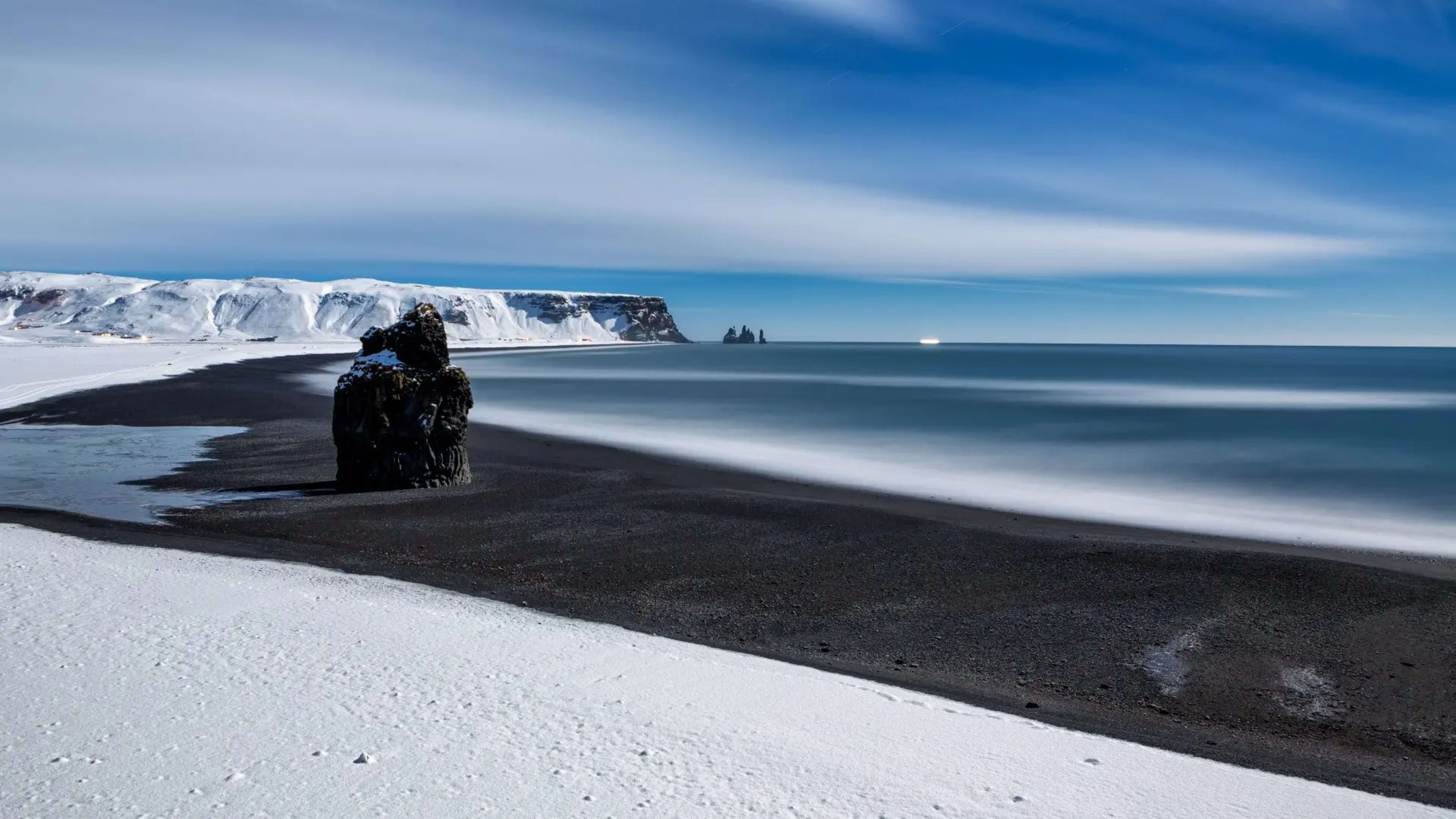 rock formation on black sand beach with layer of snow and blue sky