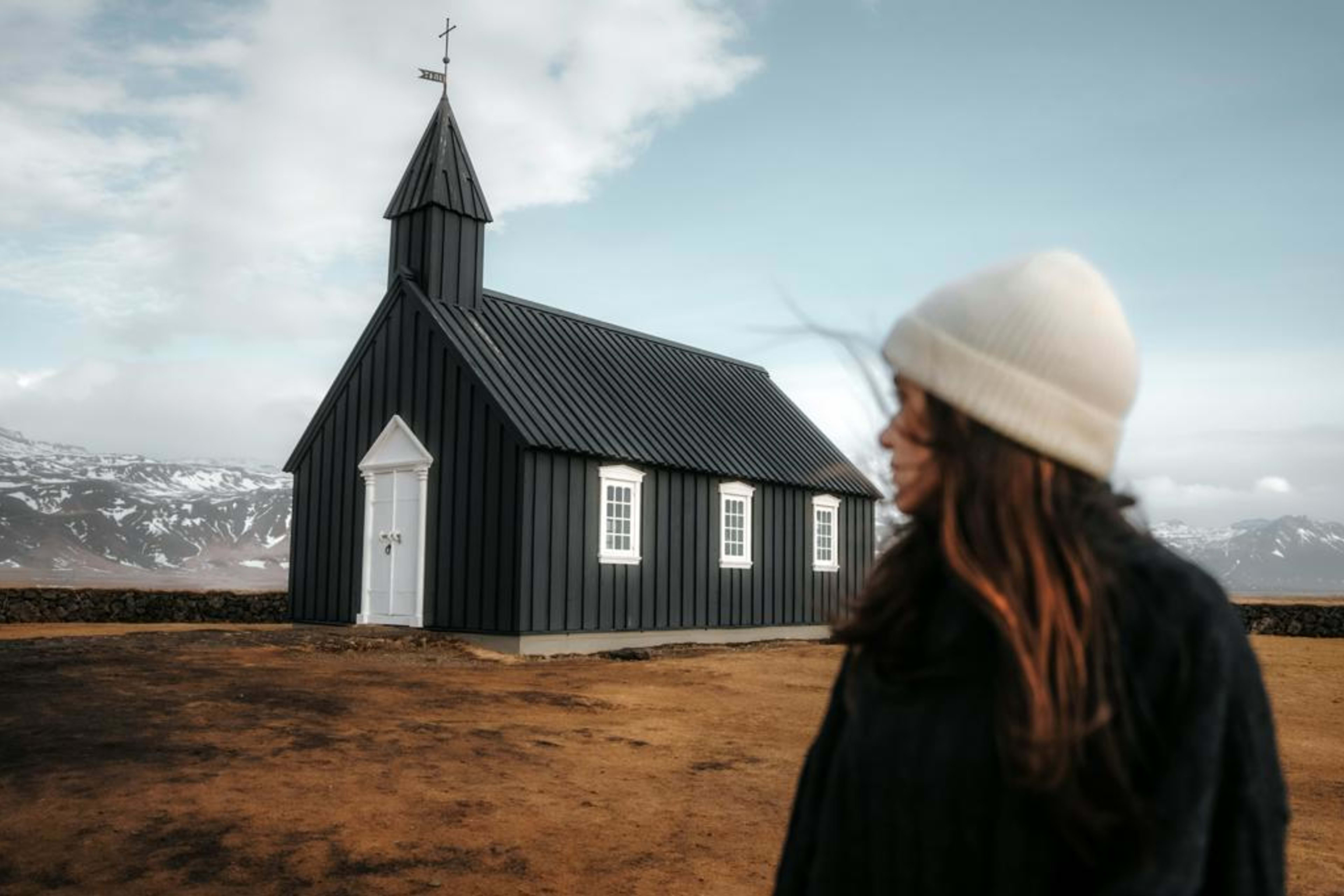 : Woman in a white hat looking towards the black wooden Búðakirkja church on the Snæfellsnes Peninsula, Iceland.