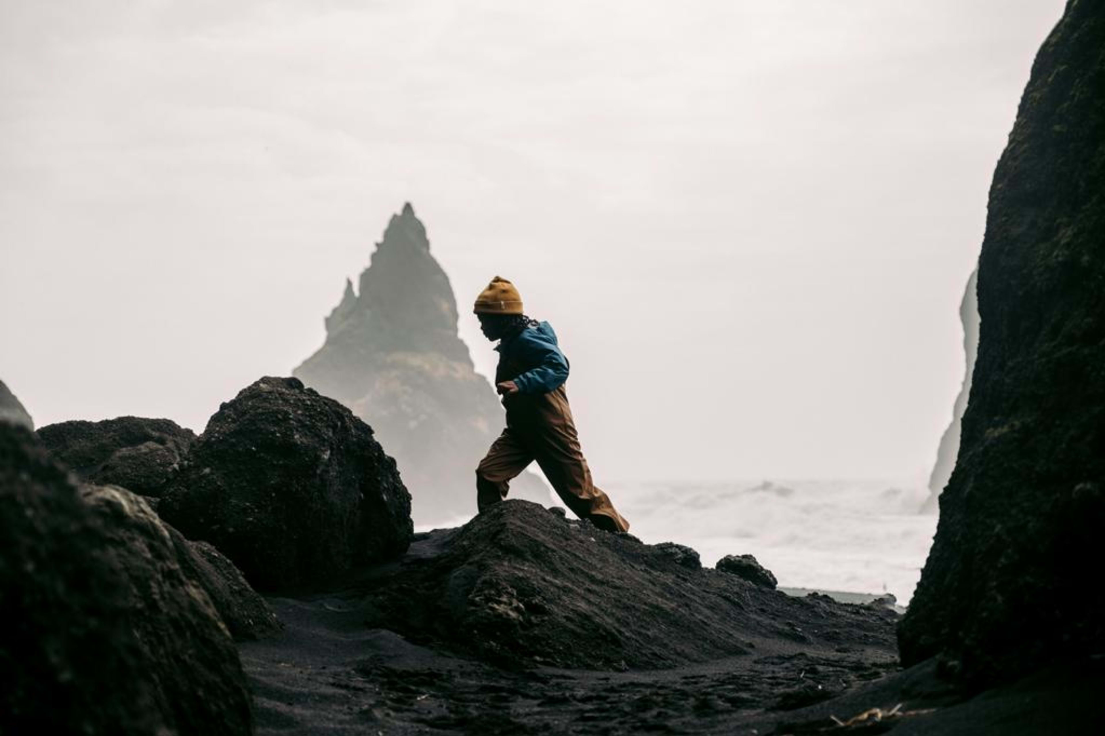 A hiker in a yellow jacket climbs between dark volcanic rocks overlooking the ocean and a pointed sea stack.