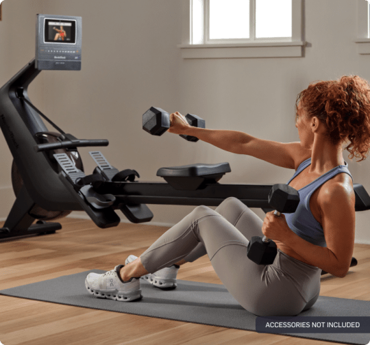 woman using dumbbells on an exercise mat next to the nordictrack rw600, while an ifit trainer is leading a workout on the console