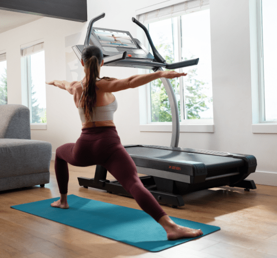 Woman stretching next to her treadmill in a lunge position, all from the comfort of her living room.