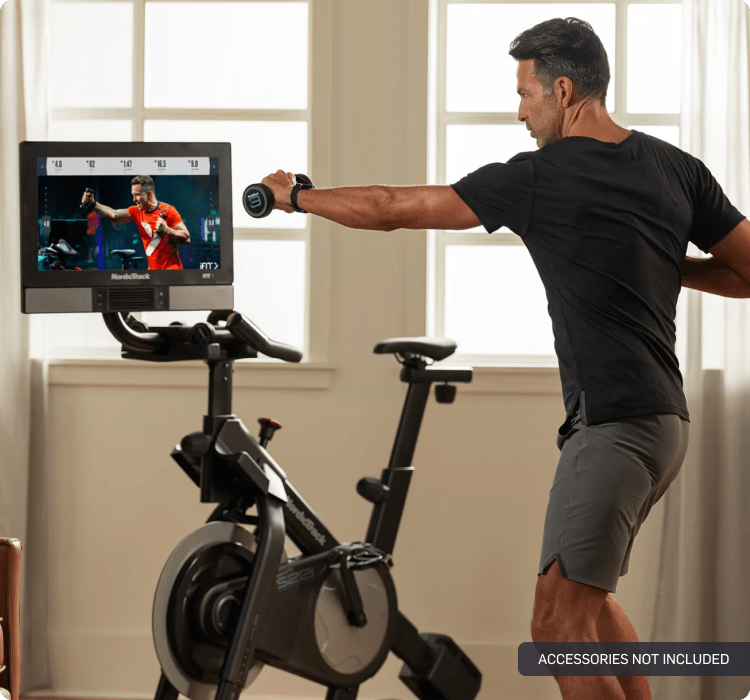 Man off of his bike, looking at iFIT trainer on a tilted screen. one hand is gripping a dumbbell and arm is extended