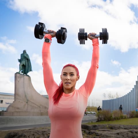 Trainer raising dumbbells high above her head outside with clouds in the background.