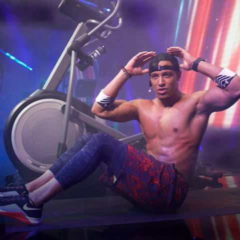 Shirtless Trainer doing sit-ups with bulging muscles and hands behind his head, facing the camera.